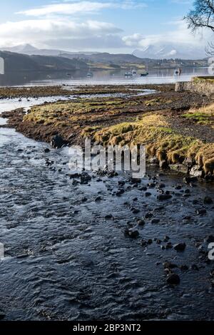 Un ruscello conduce al porto di Portree, la capitale dell'Isola di Skye in una giornata tranquilla in primavera con montagne innevate in lontananza. Foto Stock