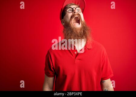 Giovane bel delivery uomo che indossa occhiali e berretto rosso su sfondo isolato arrabbiato e pazzo urlando frustrato e furioso, gridando con rabbia. Foto Stock
