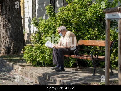 Un anziano gentiluomo che indossa una maschera a causa del virus COVID-19 sta leggendo un libro sulla passeggiata sotto il sole primaverile. Quartiere del castello di Buda, Budapest Foto Stock