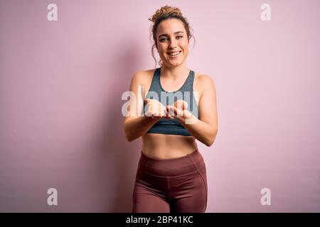 Giovane bella sportiva bionda che fa sport con indossarie sportive su sfondo rosa sorridendo con le mani palme insieme che ricevono o danno un gesto. Foto Stock