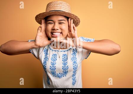 Giovane bella ragazza asiatica che indossa t-shirt casual e cappello in piedi su sfondo giallo sorridente allegro giocando a peek un boo con le mani che mostrano il volto. Foto Stock