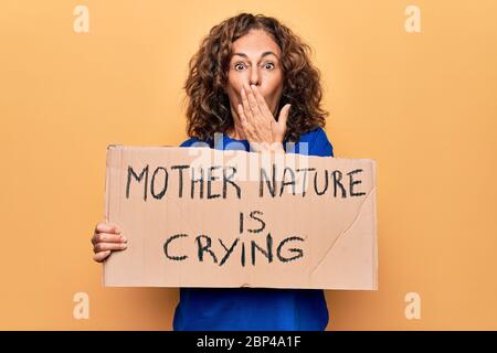 Donna di mezza età che chiede l'ambiente che tiene bandiera con la natura madre sta piangendo messaggio che copre la bocca con la mano, scioccata e impaurita per errore. Foto Stock