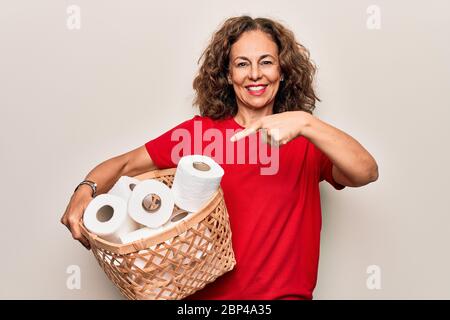 Donna di mezza età che tiene il cesto di vimini con i lotti dei rotoli di carta igienica sopra lo sfondo bianco sorridente che punta felice con la mano e il dito Foto Stock