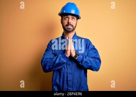 Uomo meccanico con barba che indossa uniforme blu e casco di sicurezza su sfondo giallo che prega e che prega con le mani insieme con espressione di speranza su Foto Stock