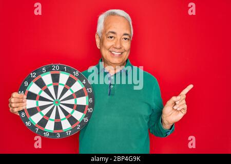 Medio età anziano uomo con capelli grigi che tiene la concorrenza bersaglio della tavola da disegno su sfondo rosso molto felice che punta con mano e dito a lato Foto Stock