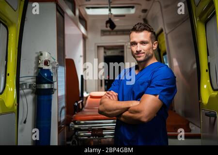 Bel paramedico in un uniforme medica guardando la macchina fotografica e sorridente, ambulanza auto in background Foto Stock