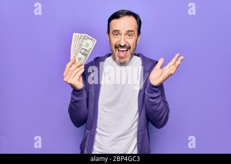 Bell'uomo di mezza età che tiene mazzo di banconote di dollari su sfondo viola isolato che celebra il successo con sorriso felice e vincitore espresso Foto Stock
