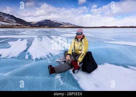 Donna sorridente con giacca gialla si siede sul ghiaccio blu del lago Baikal e cambia i suoi stivali rossi in pattini da ghiaccio Foto Stock