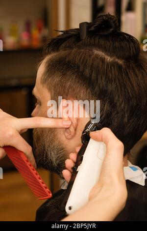 Rasoio elettrico per taglio di capelli maschile. La mano del parrucchiere rasa i capelli con il rasoio elettrico. Hipster man ottenere un taglio di capelli trendy con un regolacapelli e pettine. Servizi Barbiere. Procedura di acconciatura. Foto Stock