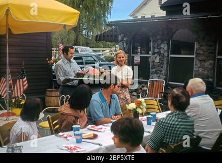 Un pasto all'aperto per famiglie, USA 1979 – una coppia porta in un intero maialino arrosto su un grande piatto. Ci sono bandiere americane nei piantatori di giardino. Sul tavolo ci sono tazze di carta e tovaglioli. Foto Stock