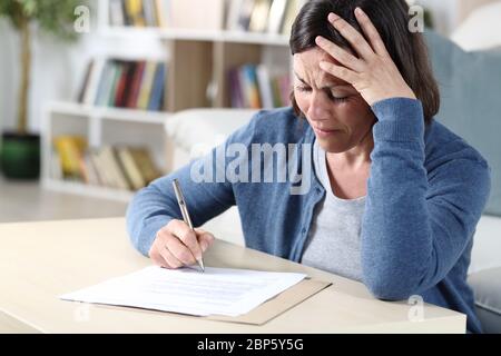 Triste donna di mezza età che firma il documento lamentando seduta nel soggiorno a casa Foto Stock