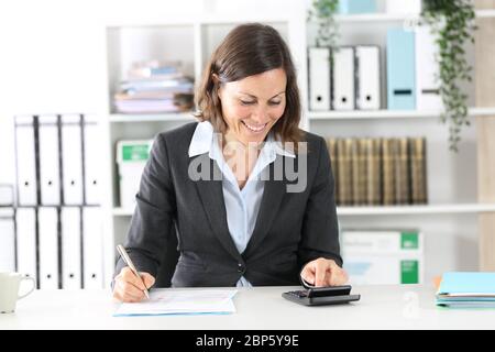 Felice donna bookkeeper adulto che calcola il saldo del conto sulla calcolatrice seduta su una scrivania in ufficio Foto Stock