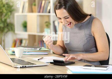 Donna contabile eccitata che calcola sulla calcolatrice che celebra il risultato seduto su una scrivania a casa Foto Stock