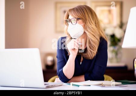 Ritratto di una donna pensante che indossa una maschera mentre si siede dietro il suo laptop e lavora da casa. Ufficio domestico. Foto Stock