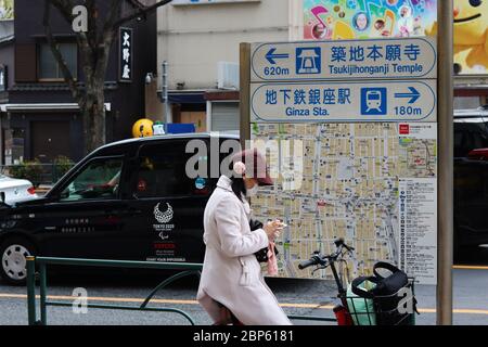 Un ciclista che indossa una maschera facciale controlla il suo smartphone sulla mappa a Ginza durante l'epidemia di coronavirus. Il taxi con il logo Paralimpico di Tokyo è sulla strada. Foto Stock