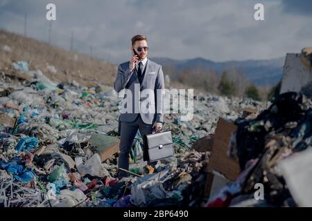 Uomo d'affari con smartphone in discarica, consumismo contro concetto di inquinamento. Foto Stock