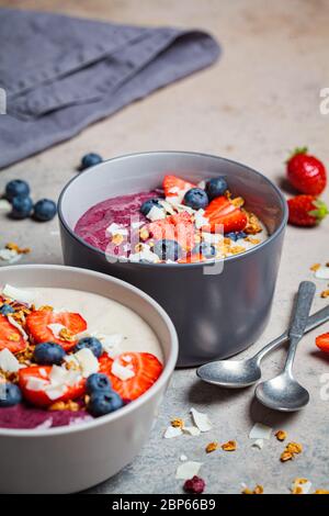 Due ciotole di frullato di frutti di bosco colorate con muesli, mirtillo e cocco, sfondo grigio. Concetto di cibo vegano sano. Foto Stock