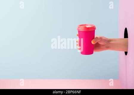 Mano femminile che tiene una tazza di caffè di carta rosa su sfondo blu e rosa Foto Stock