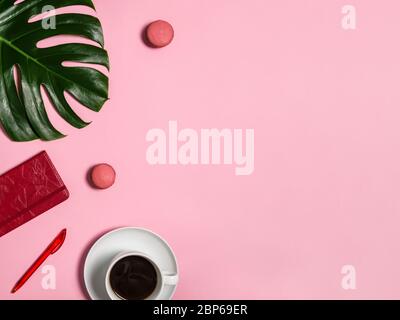 Spazio di lavoro femminile con taccuino e penna rossi, macaron, tazza bianca di caffè nero e foglia verde di monstera su sfondo rosa. Ufficio, agenda o piano di casa Foto Stock