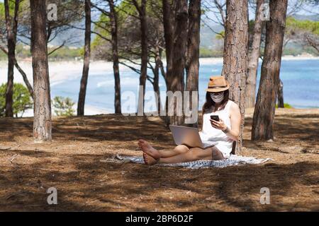 Donna freelance che lavora in vacanza sul computer portatile seduto in una pineta sul mare con maschera di protezione per coronavirus, intelligente vita di lavoro Foto Stock