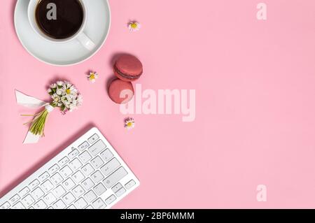 Spazio di lavoro femminile con tastiera, tazza di caffè nero, due macaron e mazzo di fiori margherita su sfondo rosa dall'alto. Sede, freelance o. Foto Stock