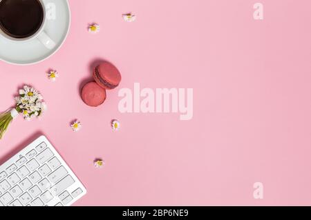 Spazio di lavoro femminile con tastiera, tazza di caffè nero, due macaron e mazzo di fiori margherita su sfondo rosa dall'alto. Sede, freelance o. Foto Stock