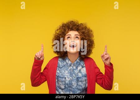 Pubblicità incredibile qui! Ritratto di donna con capelli ricci stupiti che punta in su e guarda con bocca aperta, espressione scioccata, mostrando spazio per la copia Foto Stock