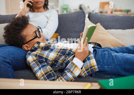 Ritratto di un ragazzo africano carino con grandi occhiali da lettura mentre si sdraia sul divano con il concetto di mamma, homeschooling Foto Stock