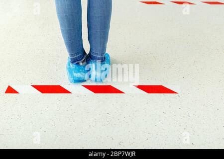 Donna stare in linea mantenendo la distanza sociale indossare copriscarpe, copriscarpe mediche in ospedale. Persone che si trovano dietro una linea di avvertimento durante la covid 19 Foto Stock