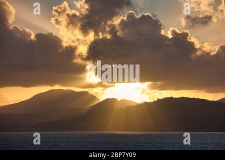 Alba sulla costa occidentale di Santa Lucia, Caraibi, Indie Occidentali, con colline in silhouette e raggi di sole luminoso contro le drammatiche nuvole scure Foto Stock