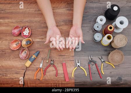 Mani di donna che mostrano nuovi orecchini che ha fatto con attrezzi, fili e perline intorno, vista dall'alto Foto Stock