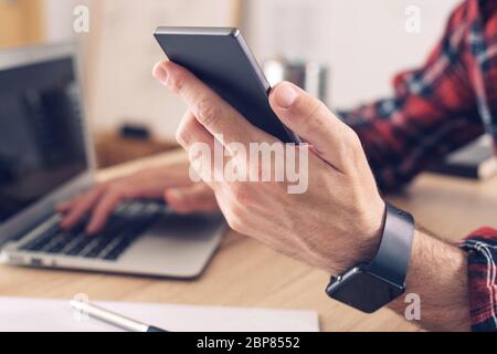 Utilizzando smartphone e computer portatile in ufficio, primo piano di mani maschili utilizzando elettronica moderna per il telelavoro, messa a fuoco selettiva Foto Stock