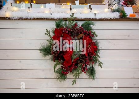 Una bella corona natalizia di rami di abete conifere, bacche rosse e fiori in brina appesa sullo sfondo di un bancone di legno chiaro Foto Stock