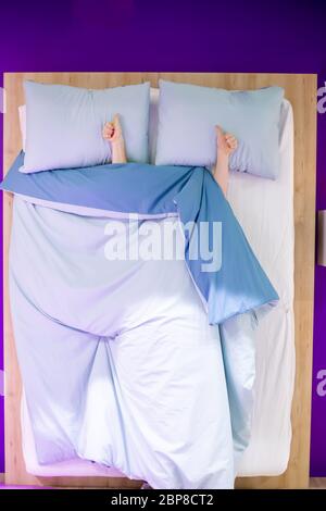 Segno di come pollici in su mostrano due mani femminili che si trovano su un cuscino da sotto le coperture blu nel letto, vista superiore, letto di legno Foto Stock