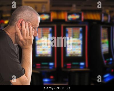 Uomo caucasico triste e depresso seduto con le mani sulla testa davanti alle file di slot machine del casinò. Immagine del tema dipendenza dal gioco d'azzardo. Primo piano Foto Stock