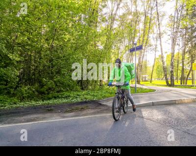 Mosca, Russia. 15 maggio 2020. Corriere in abiti verdi e in una maschera protettiva nera su una bicicletta in un parco cittadino in una giornata di sole. Consegna di cibo in qua Foto Stock