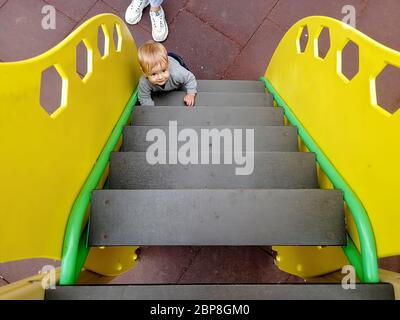 Un piccolo simpatico bambino caucasico sale le scale gialle su un parco giochi per bambini. Sul pavimento su una coperta di sicurezza gommata è la madre del bambino Foto Stock