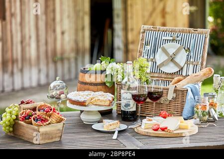 Gustoso picnic estivo all'aperto su un tavolo da giardino con vino rosso, frutti di bosco freschi e frutta, piatto di formaggi, baguette, sottaceti e torta per dessert Foto Stock