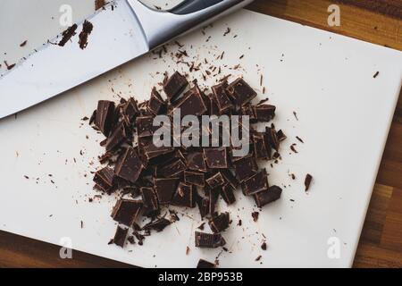 Una vista dall'alto del cioccolato tagliato a cubetti ruvidi su un tagliere bianco. Il coltello utilizzato per tagliare il cioccolato è anche in cornice Foto Stock