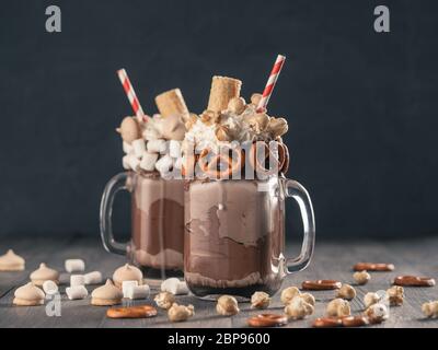 Vista ravvicinata dei due freakshake a mason jar sul tavolo marrone. Freaked frullato al cioccolato, pretzel, marshmallow, popcorn e waffles. Cibo alla moda Foto Stock