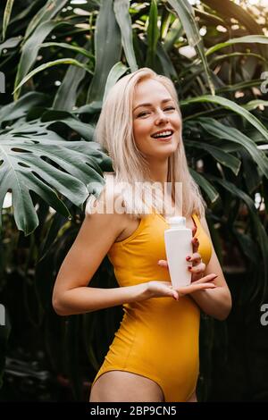 Splendida donna bionda caucasica con pelle perfettamente pulita, in costume da bagno giallo con prodotto cosmetico, bottiglia di crema solare, in piedi tra tropi Foto Stock