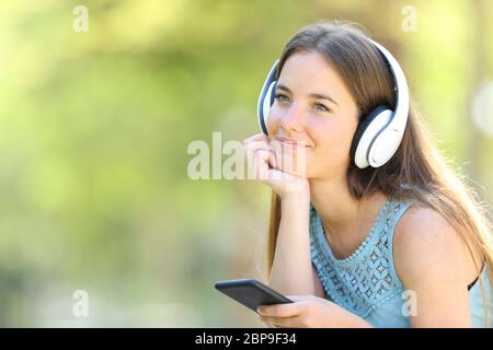 Donna rilassati ascoltando la musica azienda smart phone e guardando lontano in un parco con uno sfondo verde Foto Stock