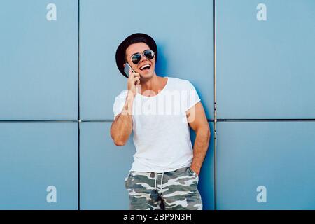 Ridendo giovane uomo parla al telefono cellulare, indossare un cappello nero e occhiali da sole, mantenendo la mano in tasca. All'esterno. Foto Stock