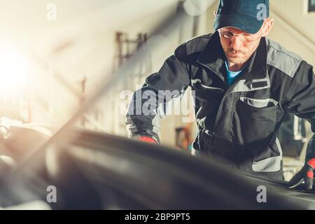 Il meccanico di automobile maschio caucasico esamina il motore e altri componenti del veicolo sotto il cofano. Foto Stock