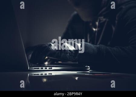 Masked Male Hacker seduto nella stanza oscura al computer portatile che commette crimini informatici. Foto Stock
