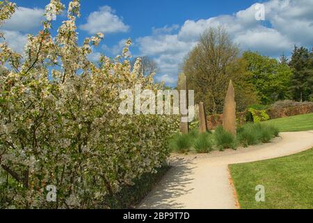 Garden Path fiancheggiato da fiori di mela bianca che portano a sculture alte e sottili in pietra scolpita, RHS Garden, Harlow Carr, Harrogate, North Yorkshire, Inghilterra. Foto Stock