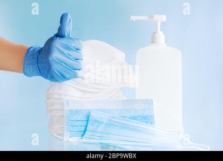 La mano del medico in guanti blu mostra i pollici sul segno. Molte maschere mediche diverse, maschera chirurgica monouso protettiva, gel igienizzante per alcool Foto Stock