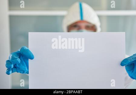 Il lavoratore di controllo delle malattie in indumenti protettivi tiene una scheda di avviso vuota con spazio di copia a causa della pandemia di Covid-19 Foto Stock