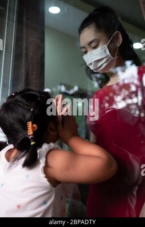 Madre indossare maschera viso incontro figlia e toccare la mano attraverso la finestra a causa della quarantena Corona Virus Covid-19, sociale di distanza Foto Stock