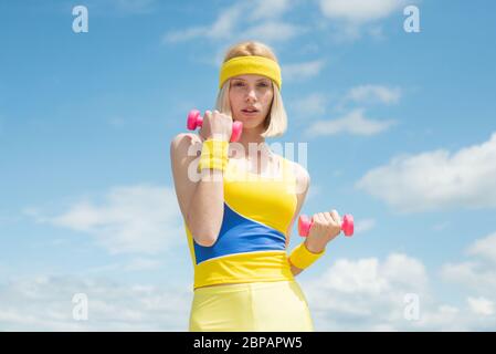 Giovane sana attraente sportivo con uniforme blu che mostra esercizi con manubri rosa su sfondo cielo con clou Foto Stock